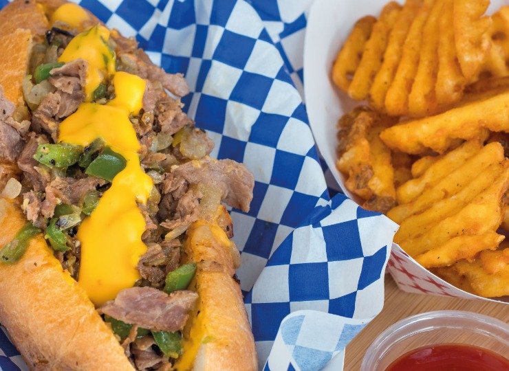 Blue checked table cloth with a philly cheese steak sub and side of waffle fries